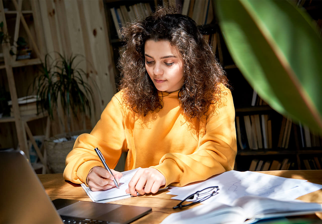 An young woman surrounded by books and a laptop takes notes using a notebook and pen.