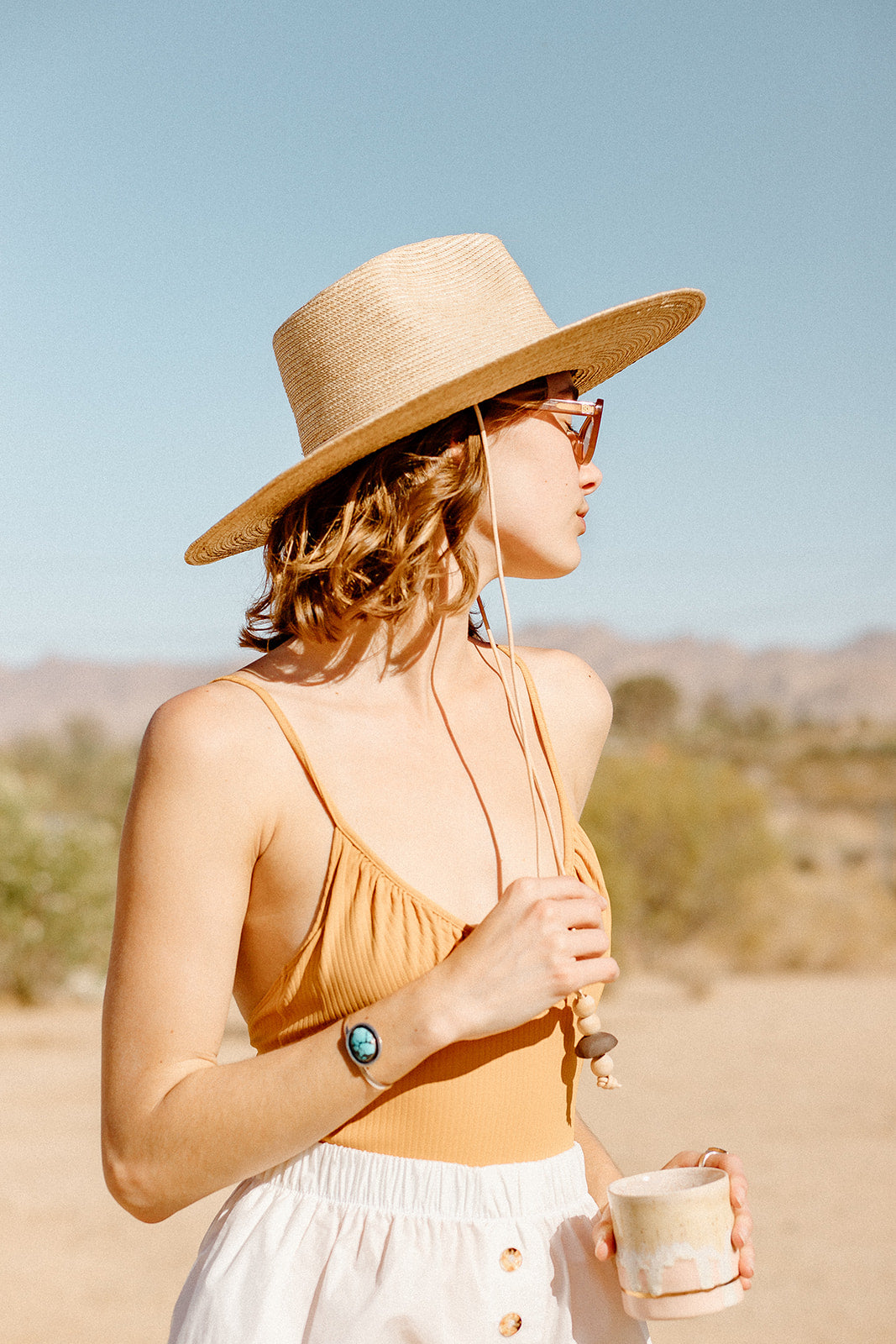 A woman wearing an ethically made one piece swimsuit with a wide brim sun hat and white shorts in the desert