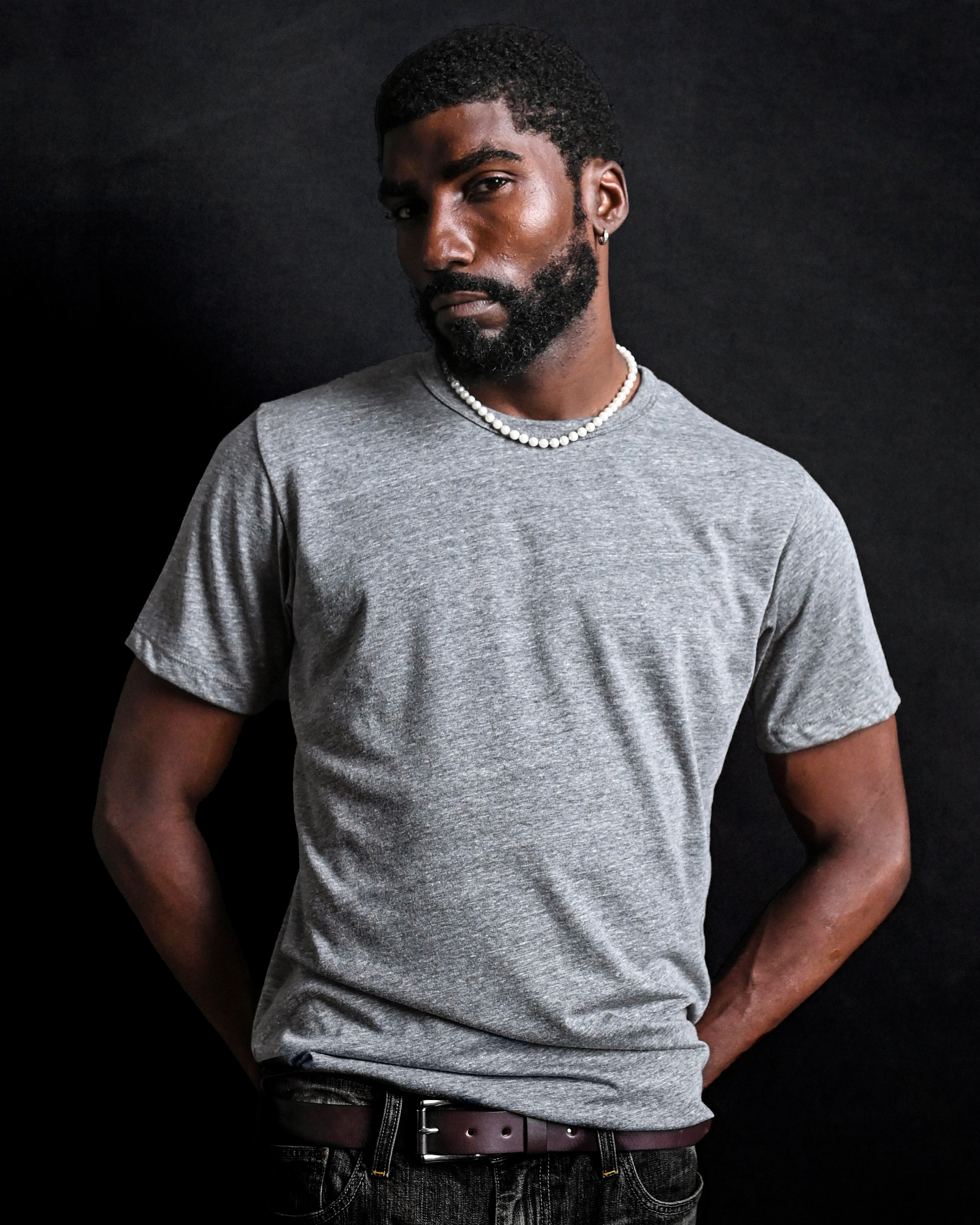 Man wearing a gray shirt and pearl necklace against a dark background.