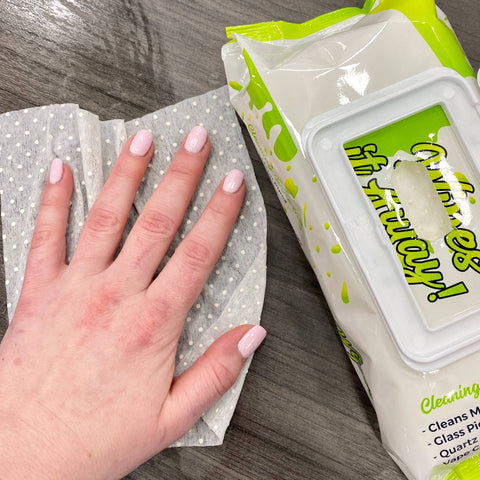 A white woman's hand with pink nails is using a Res Wipe to wipe down a table. There is a 100ct pack of wipes next to her hand.