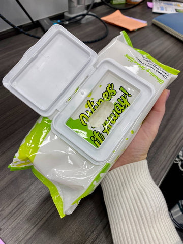 A woman wearing a white sweater is holding a 100ct pack of Ooze Resolution Wipes. The lid of the flexible packaging is open to show the top wipe.