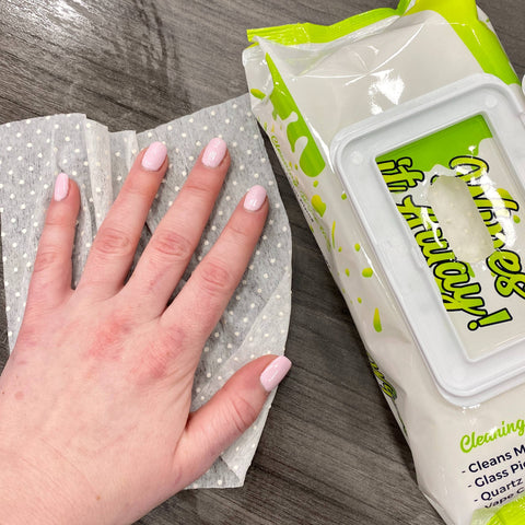 A white woman's hand with light pink nails cleans a gray wood table off with a Res Wipe. There is a 100ct pack of wipes next to her hand.