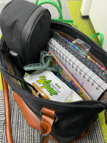 A pack of Ooze Resolution Res Wipes is in a black and brown leather travel bag with a few notebooks, a pack of markers, a hair clip, and a toiletry bag.