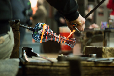 A glass artist is working on a blown glass vase water pipe piece. It is angled towards the table, and they are wrapping a bright orange hot, molten string of glass around the piece to stripe it.