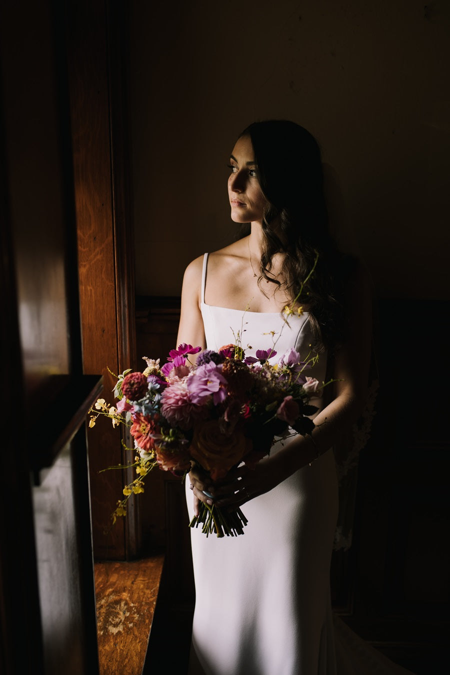 A dark and moody photo with dramatic lighting across the bride's face, front of dress, and bridal bouquet. The bouquet is full with a variety of colors.