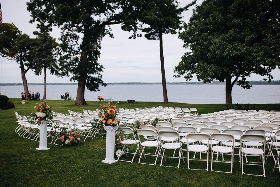 Shot of the ceremony set up. White folding chairs facing the water. Floral arrangements line the aisle on the ground and on pedestals at the top of the aisle.