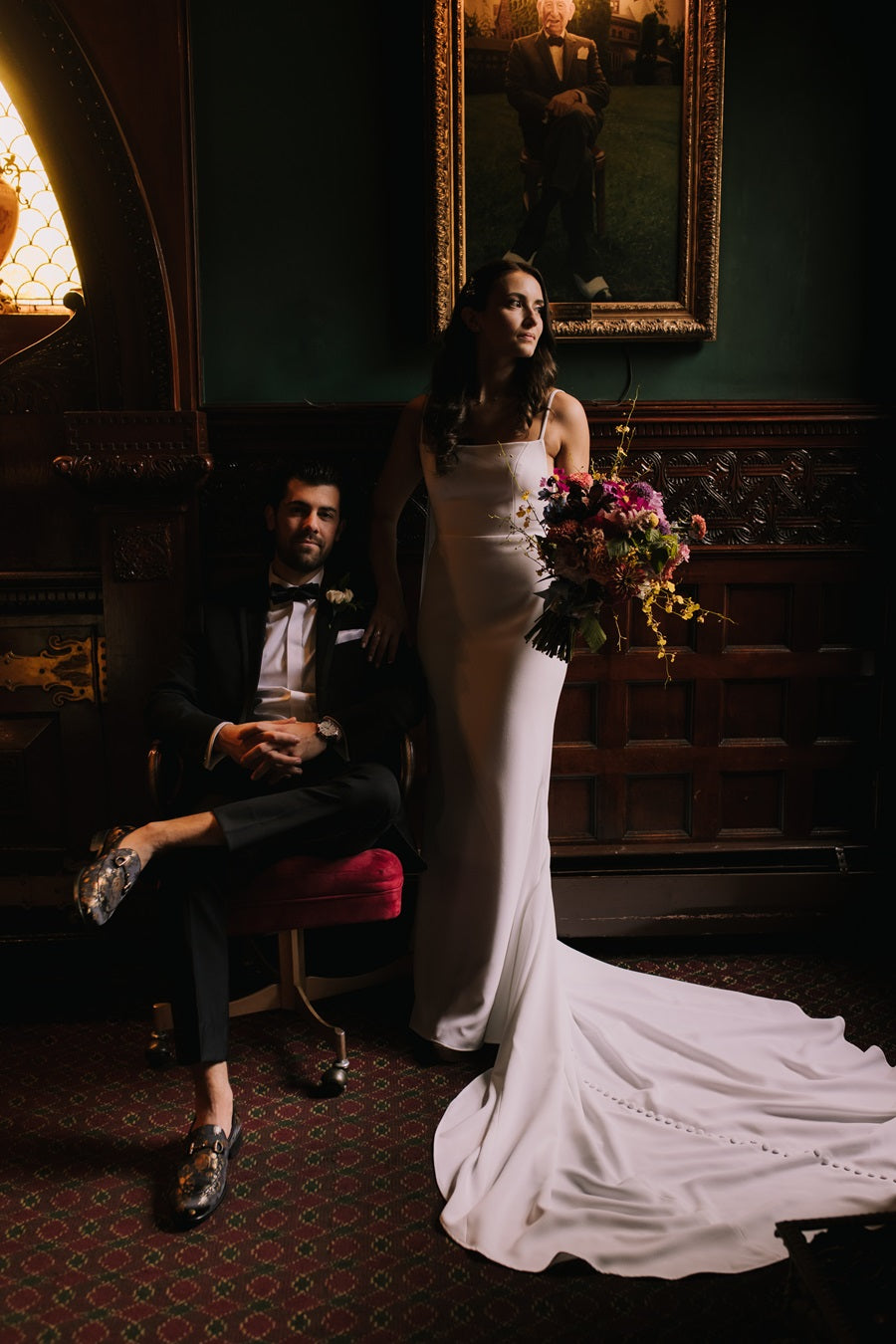 A dramatic shot featuring the bride and groom. Groom is sitting in a chair with the bride standing at his side holding her bridal bouquet. The lighting creates a dark and moody atmosphere, at Belhurst Castle