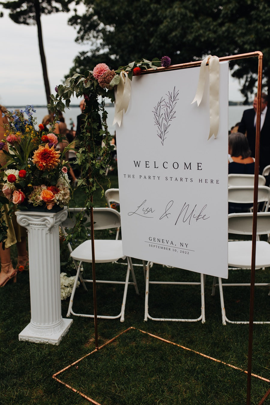 Welcome sign, white with colorful accent florals, white pedestal with bright flower arrangement.