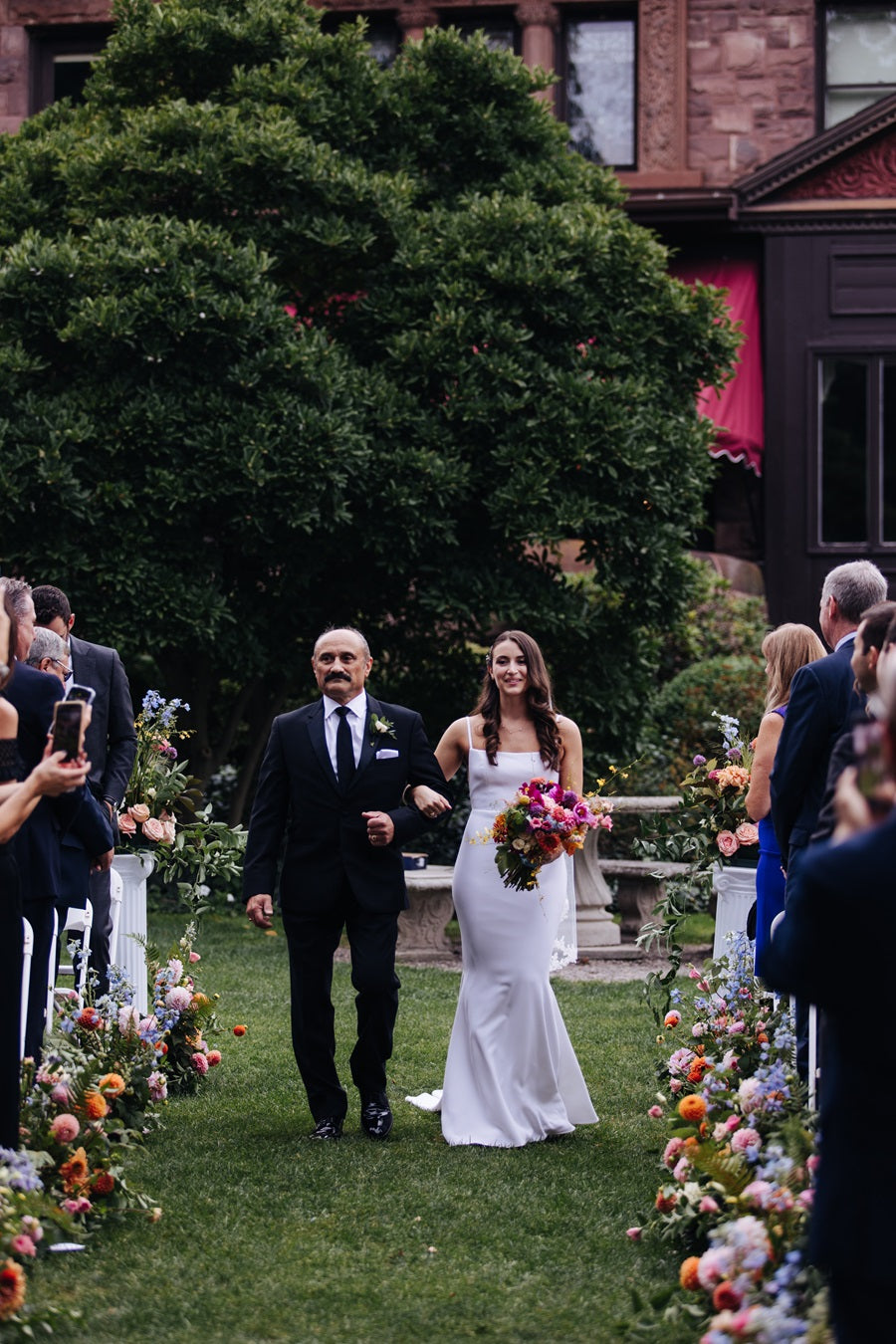 Bride being escorted down the aisle holding her bouquet. Florals line the aisle on the ground and viewers stand watching on either side.