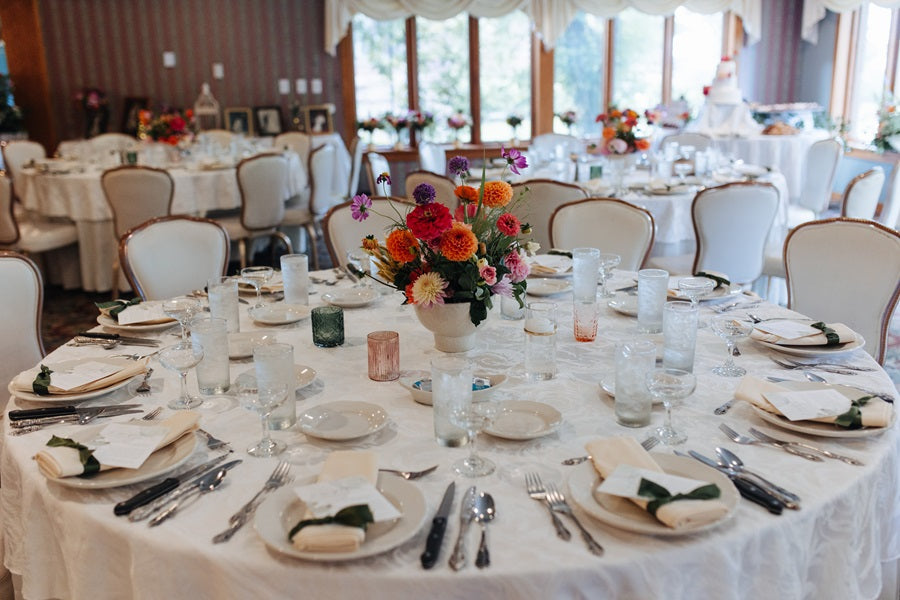 Reception table set up with neutral tableware, colorful votive candles, and a colorful floral arrangement in a white vase at the center.