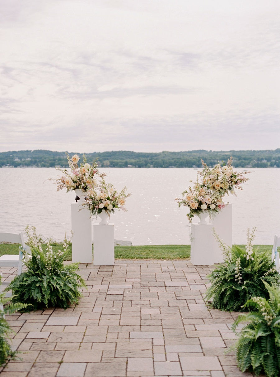 White pillars by the lake with lush floral arrangements in white, peach, pink, and green. Large fern plants with white florals line the aisle.