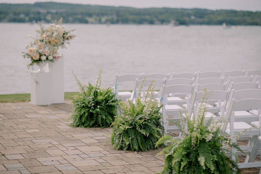 White chairs lined up facing the floral arrangements at the front. Large ferns with white florals line the sides of the aisle.