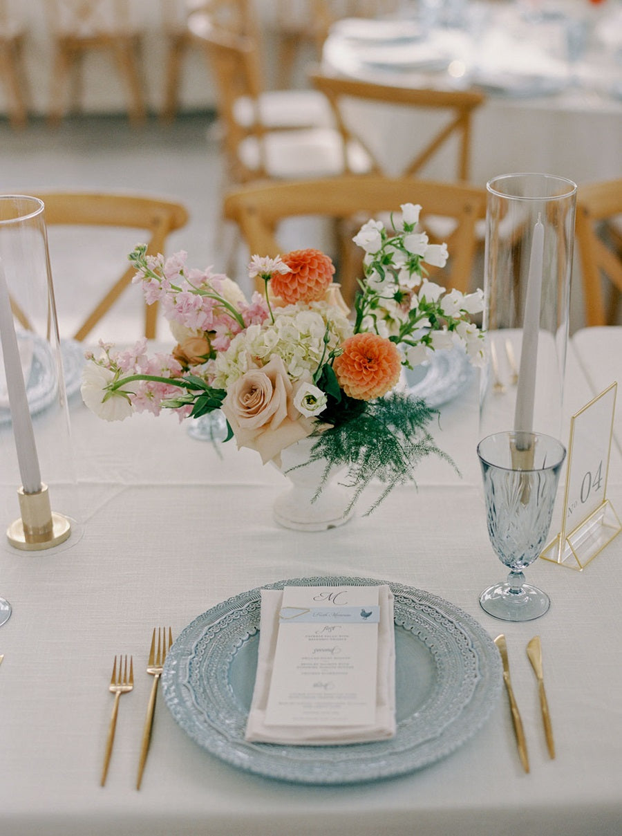 Close up on a floral centerpiece that has pink, white, orange, peach, and green. In a white compote vase. Table is set with blue and gold. Some of the flowers pictured are roses, dahlias, hydrangea, and fern.