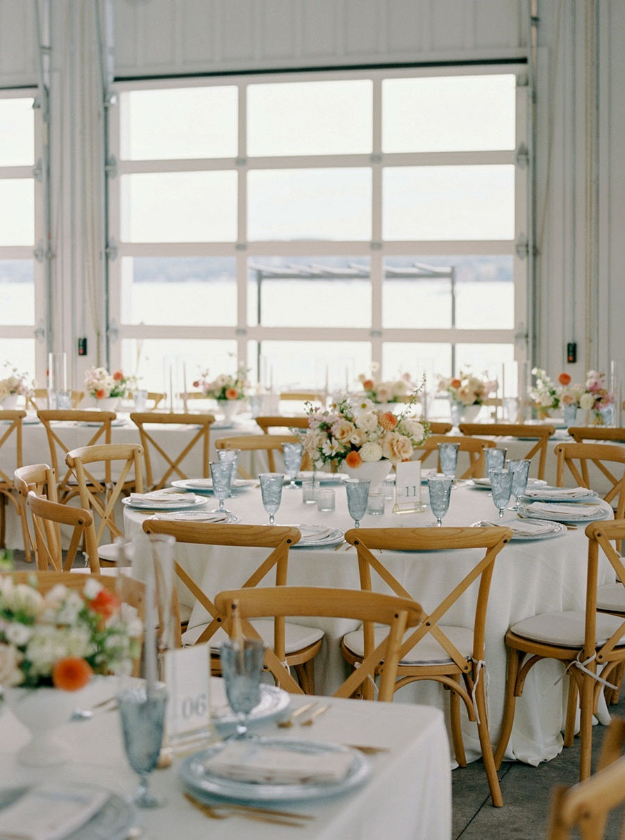 Reception shot featuring a round table. White table cloth, blue and gold tableware, and a white, peach, pink, and orange, floral arrangement.