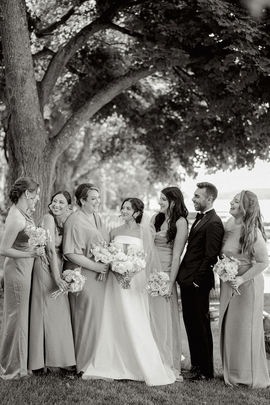 Bridal party stands with the bride. Bridesmaids hold bouquets that match the arbor pieces, and the bride holds an all white bouquet. Photo was shot in black/white.