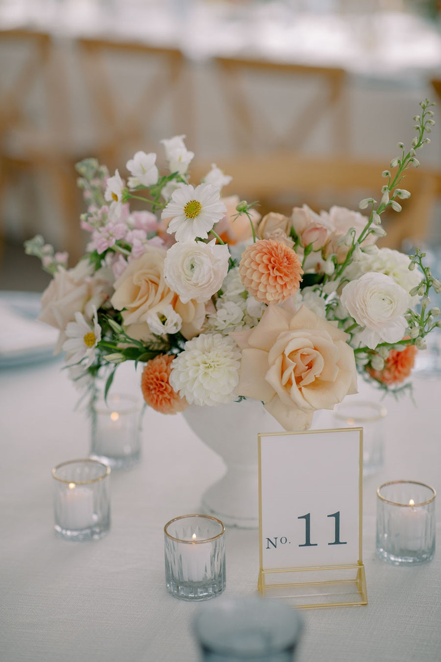 Close up on a floral centerpiece that has pink, white, orange, peach, and green. In a white compote vase. Table is set with blue and gold. Some of the flowers pictured are roses, dahlias, hydrangea, and fern.