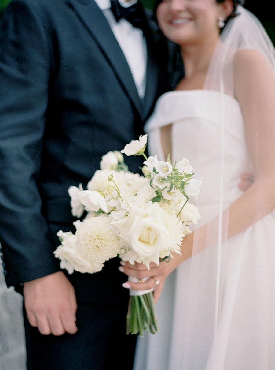 Bride and groom embraces. Close up on the bridal bouquet that is full of white flowers such as roses, dahlias, spray roses, and more.