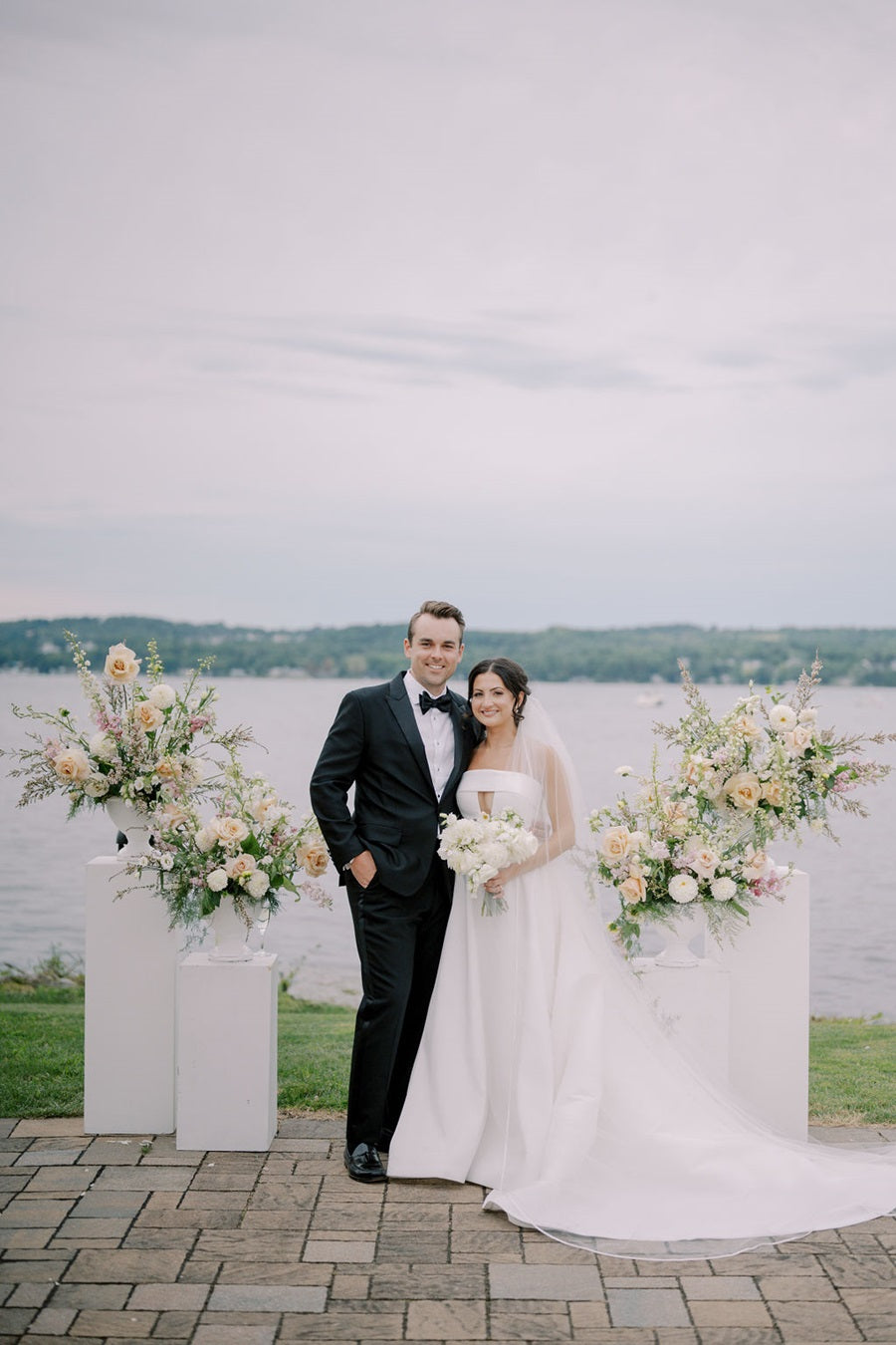 Bride and groom stand in front of the floral arrangements. Bride hold a bouquet made of all white flowers.