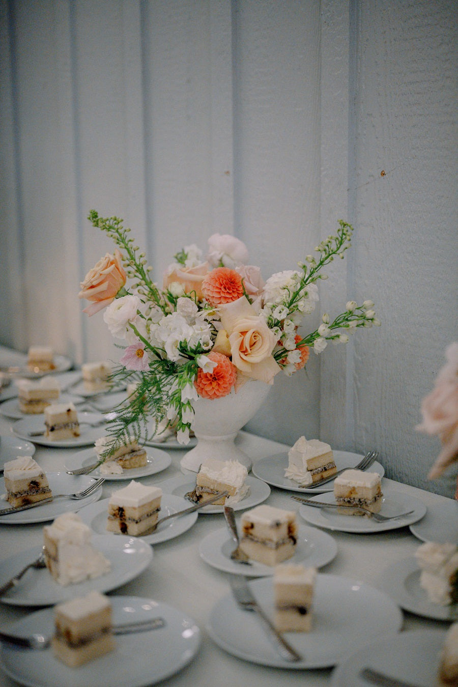 A floral arrangement with whites, peach, orange, and green, placed on the dessert table. The rest of the table is covered in places full of cake slices.