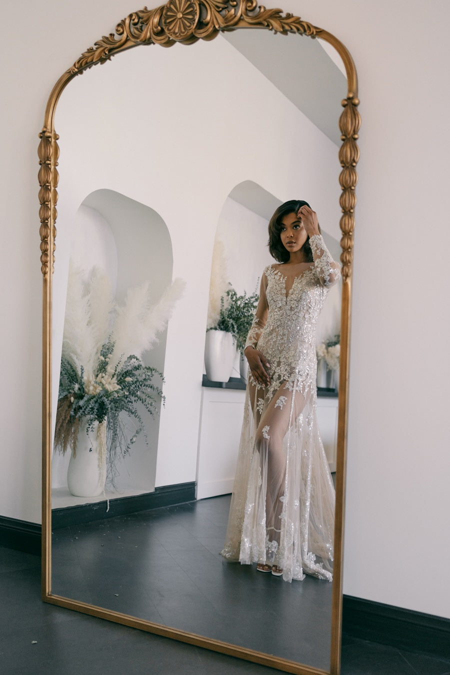 Bride stands in front of a gold standing mirror. Dried floral arrangements sit in cubbies behind her bringing in accent colors of green and beige.