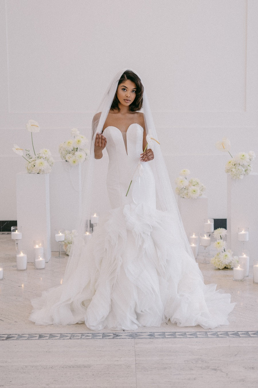 Bride in white gown with a full skirt and veil. She holds a flower in one hand and is framed by floral arrangements in the background. Lit candles are placed in sets along the base of the pedestals.