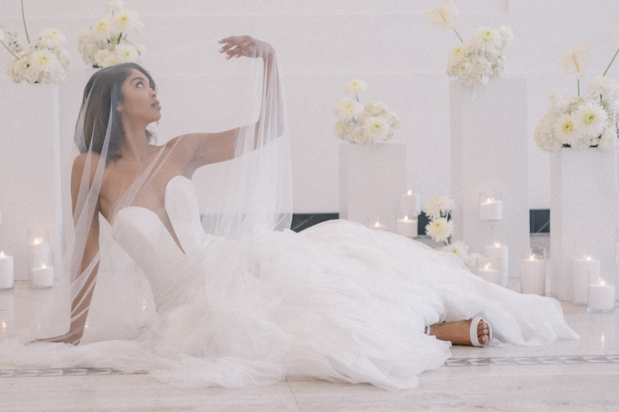 Bride lays on the ground, arm stretched upwards, covered in her veil. She is framed by floral arrangements in white pedestals with candles lining the base.