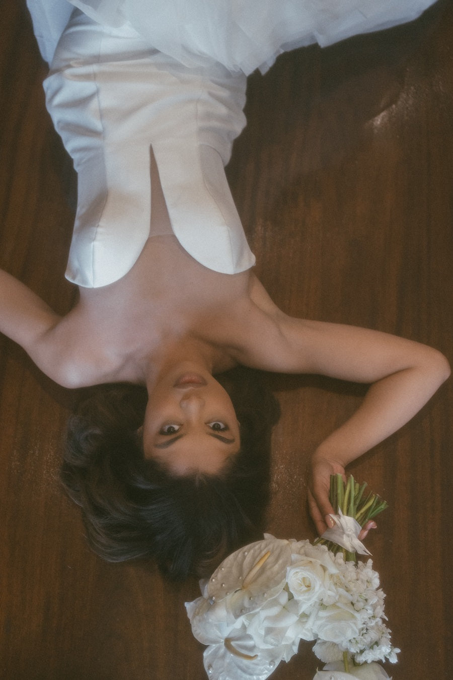 Bride lays on her back looking up at the camera. She holds a bridal bouquet of white roses and calathea.