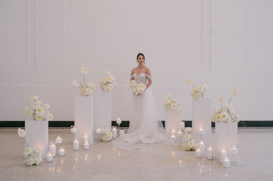 The bride stands amongst pedestals of floral arrangements and additional floral arrangements line the floor. Lit candles are placed around the base in sets. Some florals pictured are white roses and calathea. Pearl accents are placed throughout.