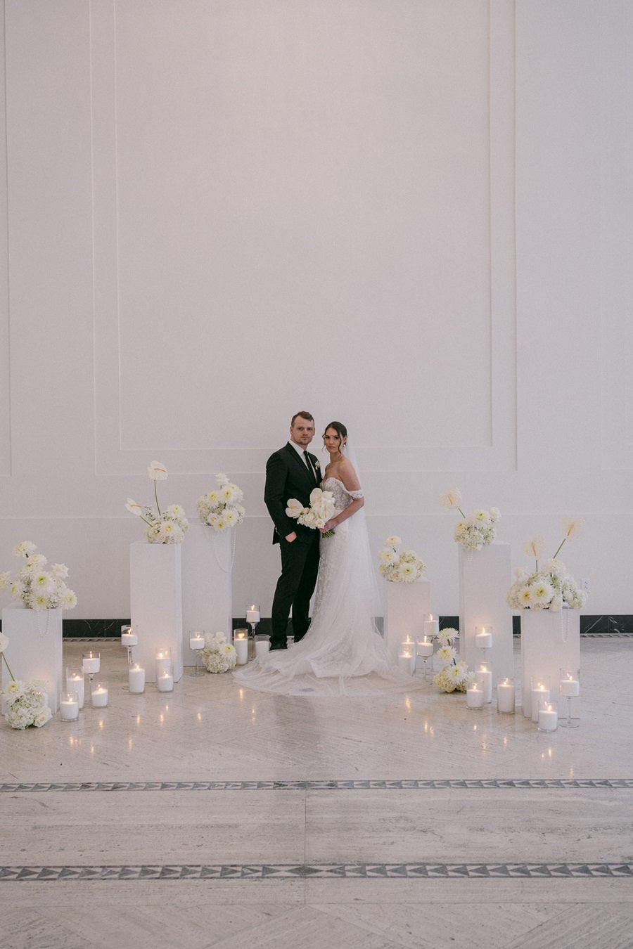 Bride and groom stand together amongst pedestals of floral arrangements and candles.