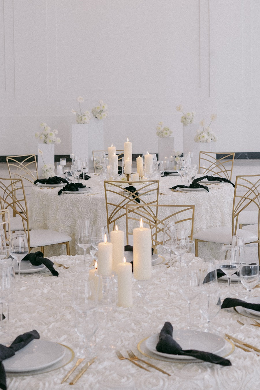 The reception tables are set in a black and white theme. The table cloths themselves are white with a textured rose pattern. The centerpieces are sets of lit pillar candles and the floral pedestals can be seen in the background.