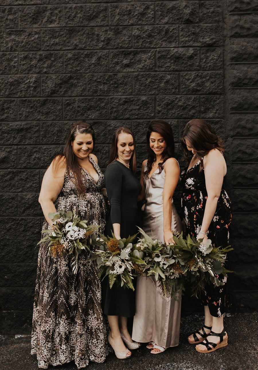 Bride poses with bridesmaids, all holding bouquets and looking down towards the ground. Black block background.