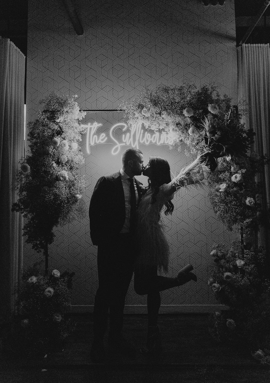 Black/White photo of bride and groom in front the a floral reception arch. The florals are lush, roses, baby's breath, and greenery. A glowing neon sign back lights the couple, bride throws her arm into the air with a popped back leg.