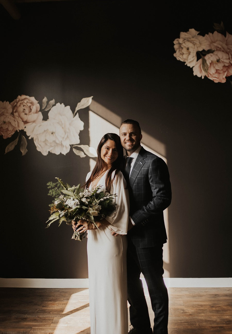 Bride and groom pose together in front of a wall with a floral design. Strong sunlight comes in from the left, highlighting the floral bridal bouquet.