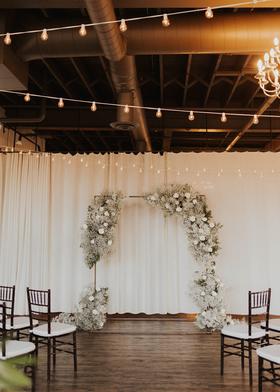 Shot of the floral arch at Arbor Loft. It is decorated with lush florals including white roses, baby's breath, and greenery. Chairs line the aisle on either side.