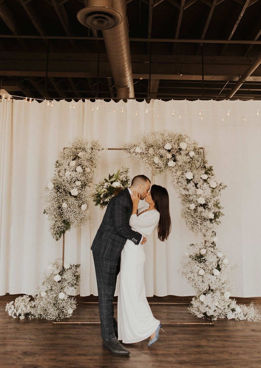 Bride and groom embrace in front of their arch. The arch is decorated with lush florals of roses, baby's breath, and greenery. Arch is placed against a curtain backdrop.