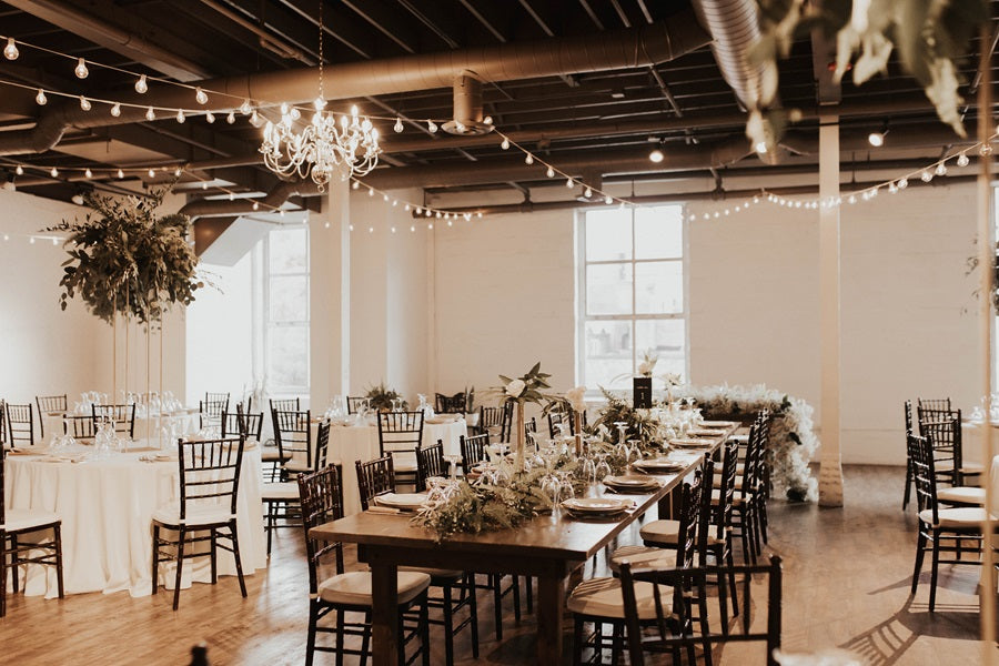 Wide room shot showing the tables, raised centerpieces, and industrial venue. The ceiling is lined with string lights.