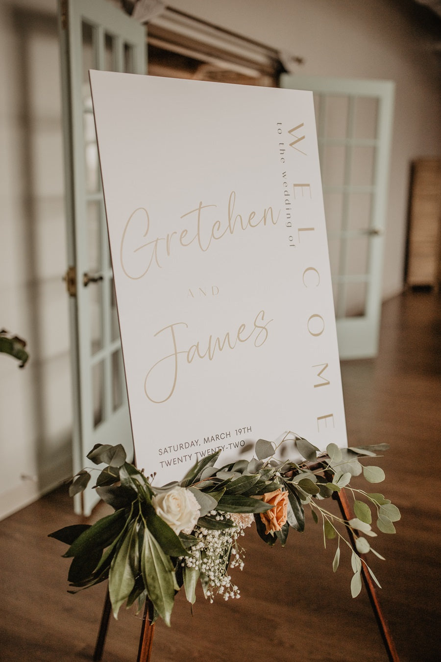 White welcome in an easel with greenery and florals at the base of the poster board.