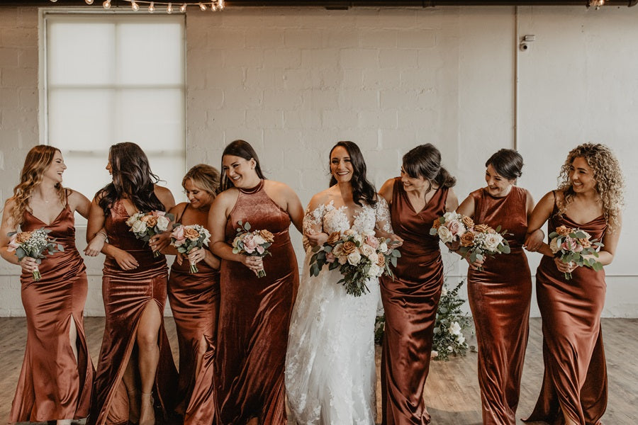 Bridal party stands together, bride in the center, all holding matching bouquets. They are dressed in a burnt/neutral orange velvet and silk in a variety of styles.