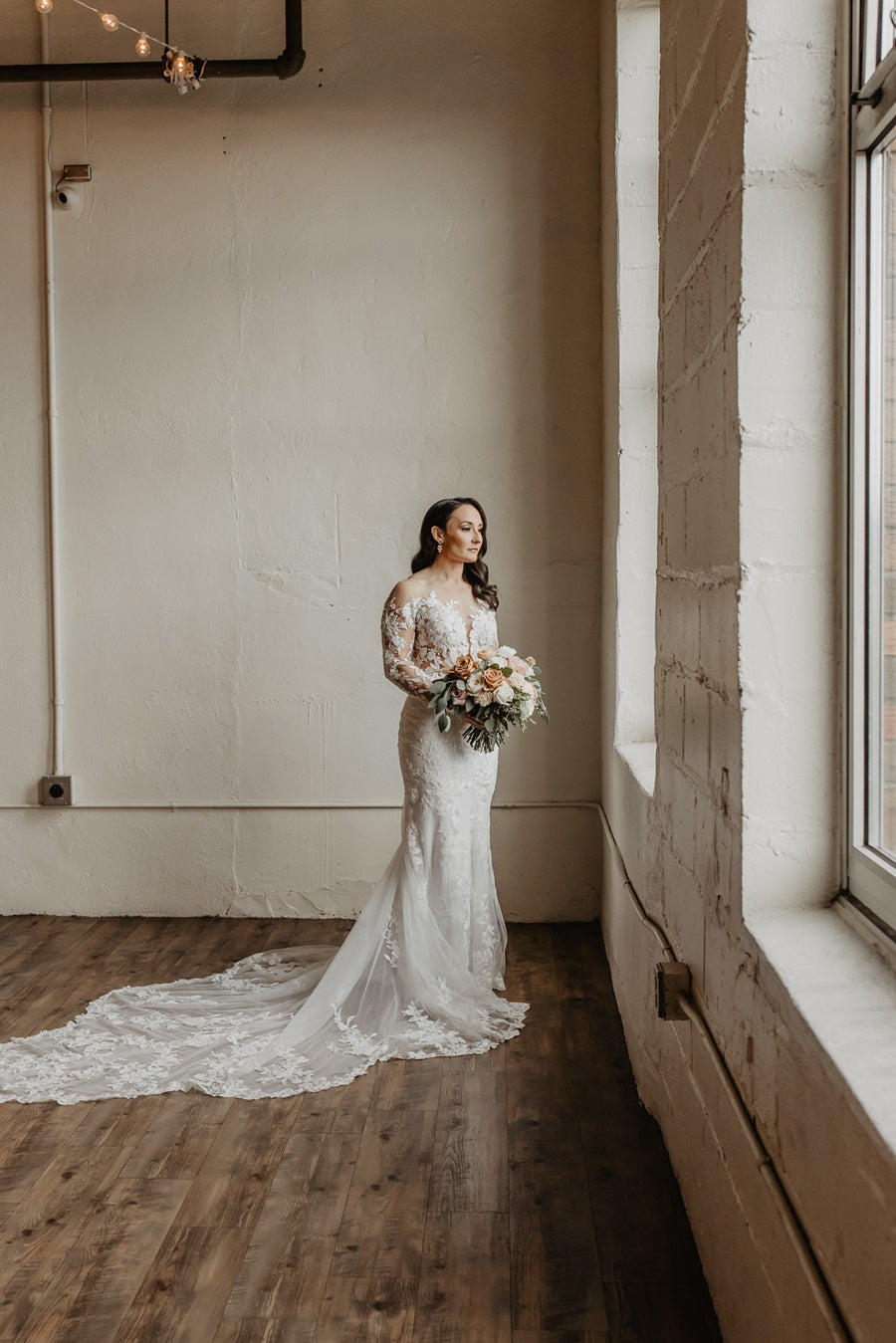 Bride in her gown holding her bridal bouquet, looking out the window.