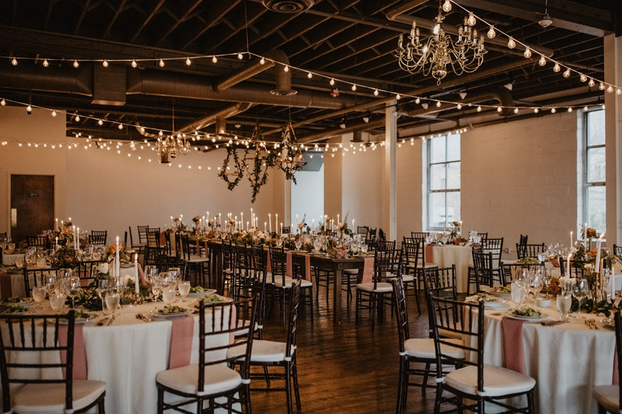 Wide shot of the room showing white linens, pink accents, greenery, taper candles, string lights, and hanging greenery.