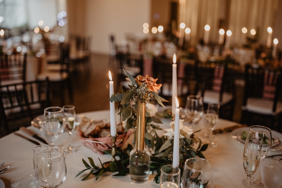 Close up on tall budvase with greenery and roses. Table is set with gold and pink accents, white taper candles, and glassware.