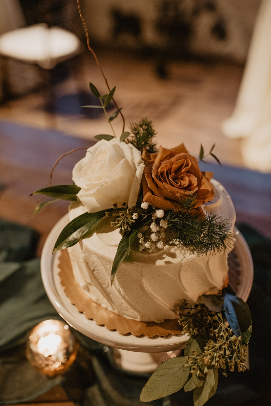 A small white cake with greenery, baby's breath, a white rose, toffee rose, and branching.