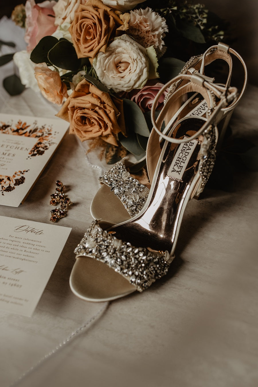 Wedding stills, shot of the bride's shows, bouquet, and wedding invitation. The bride's high heel shoes are bedazzled with silver rhinestones.