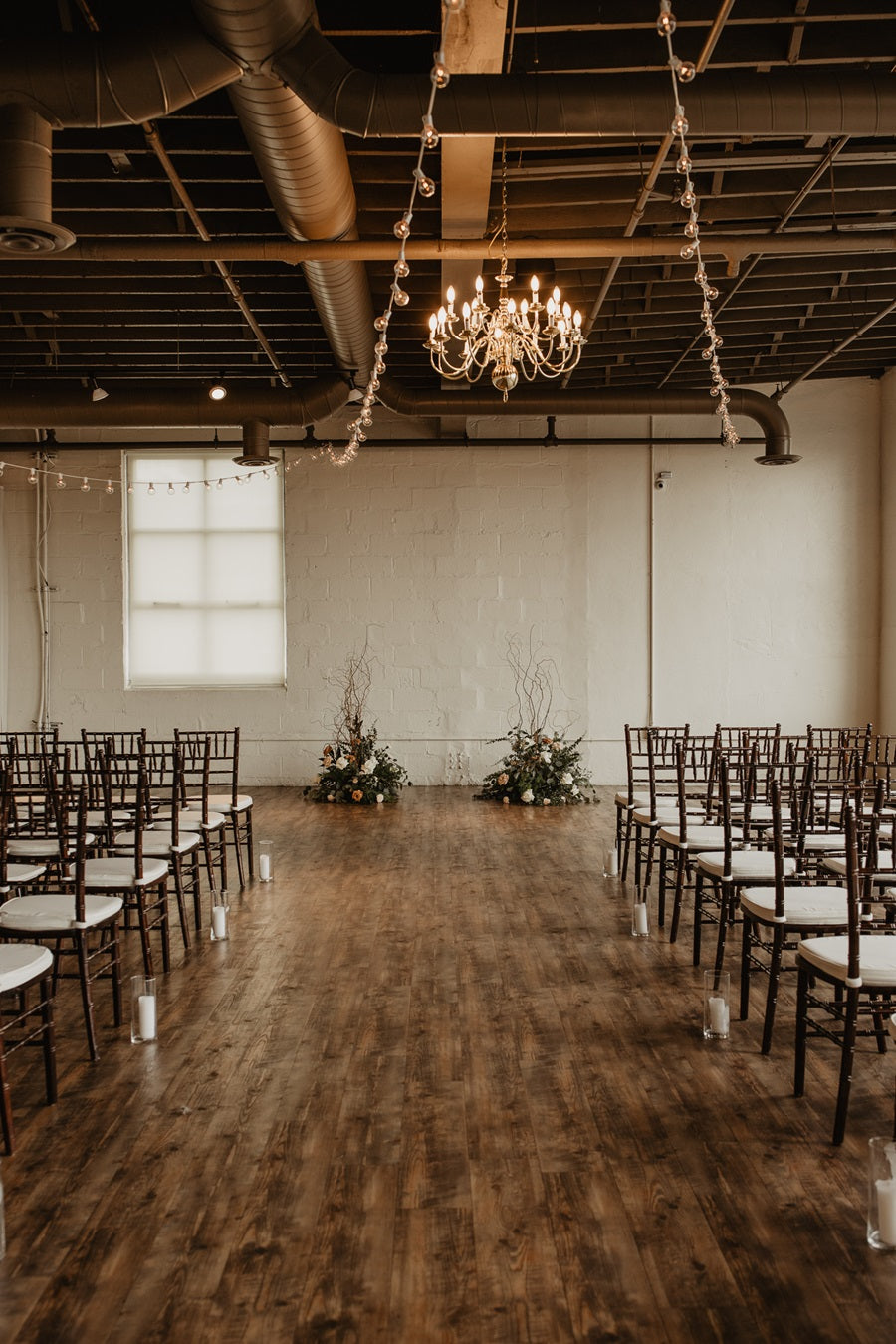 Preview ceremony shot right down the center of the aisle. Chairs line either side, with tall floral floor pieces to frame the bride and groom. Branching adds height and shape to the arrangements.