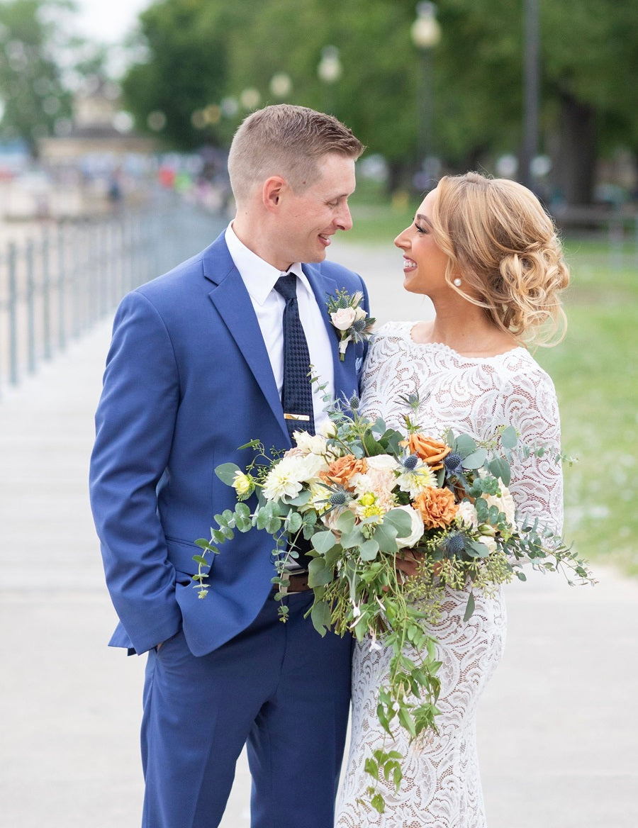 Bride in white and groom in blue, embrace on the beachside. Bride holds a bouquet of greens, whites, and blue/orange accents.