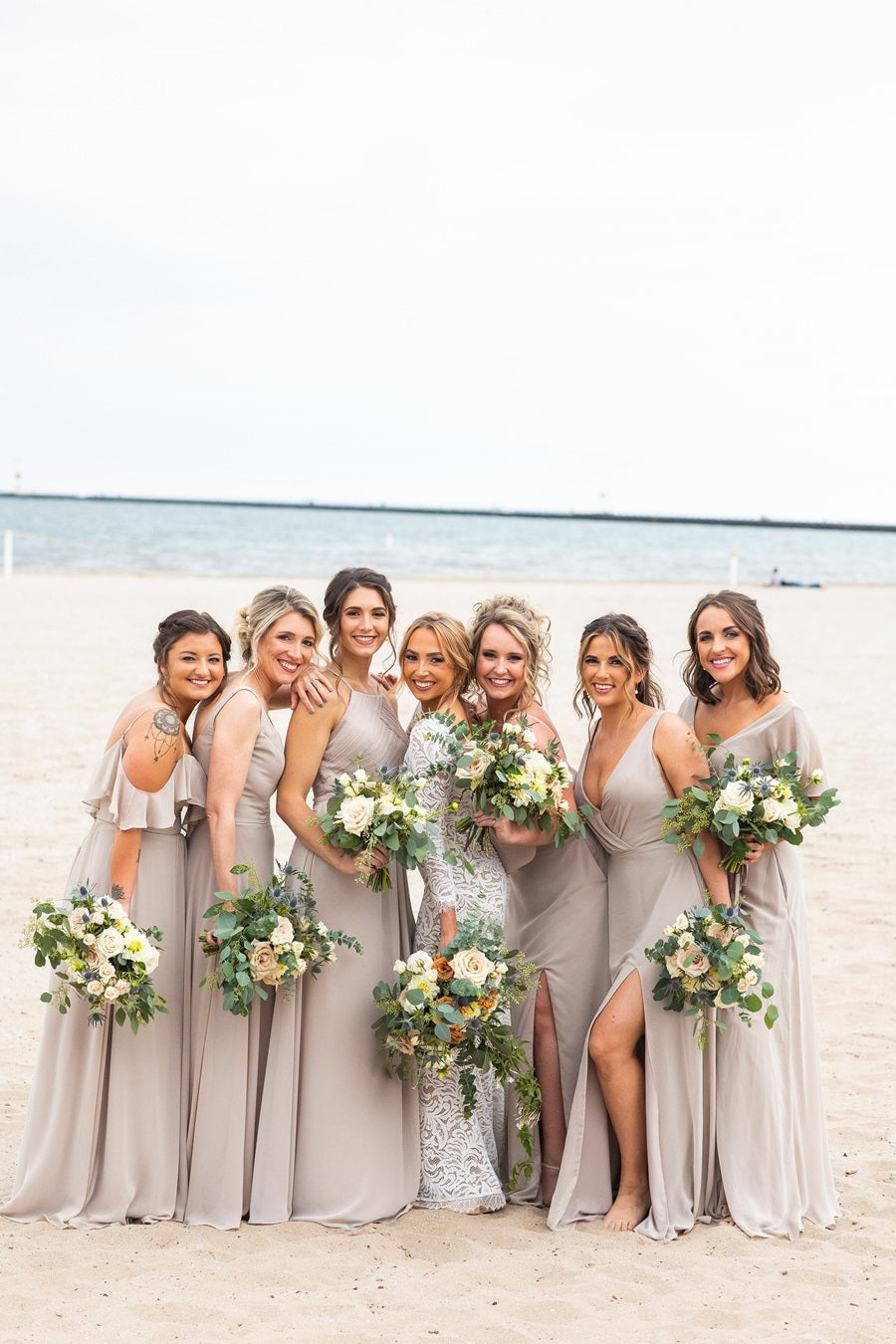 Bridal party poses on the beach, bride in white, bridesmaids in a warm light gray. All holding matching bouquets in the green, white, and blue w/ toffee accents color palette.