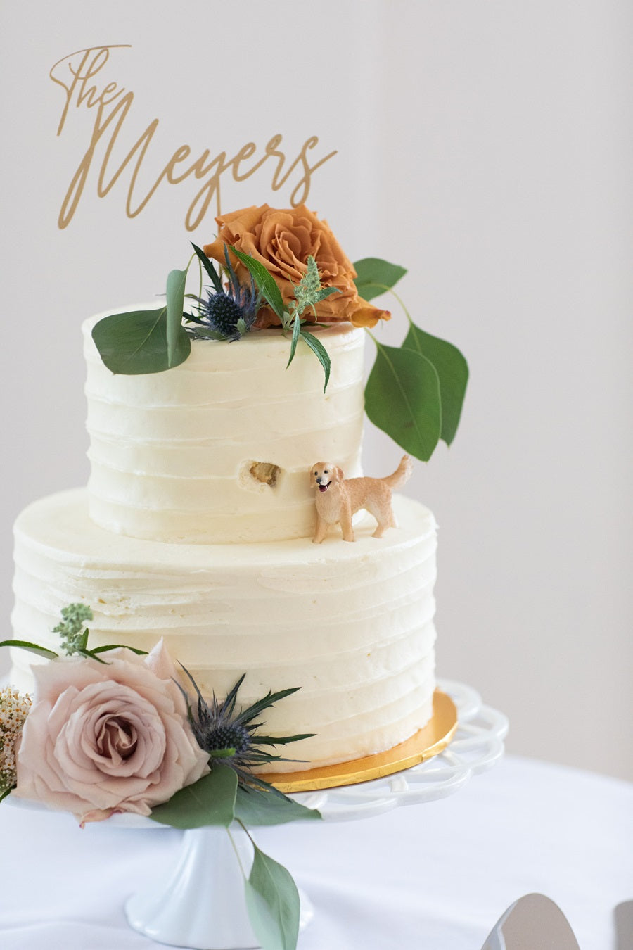 A white wedding cake with a toffee and a neutral lavender rose, accented with greenery. A dog figurine is placed on the side next to a small hole that implies it was eating the cake.