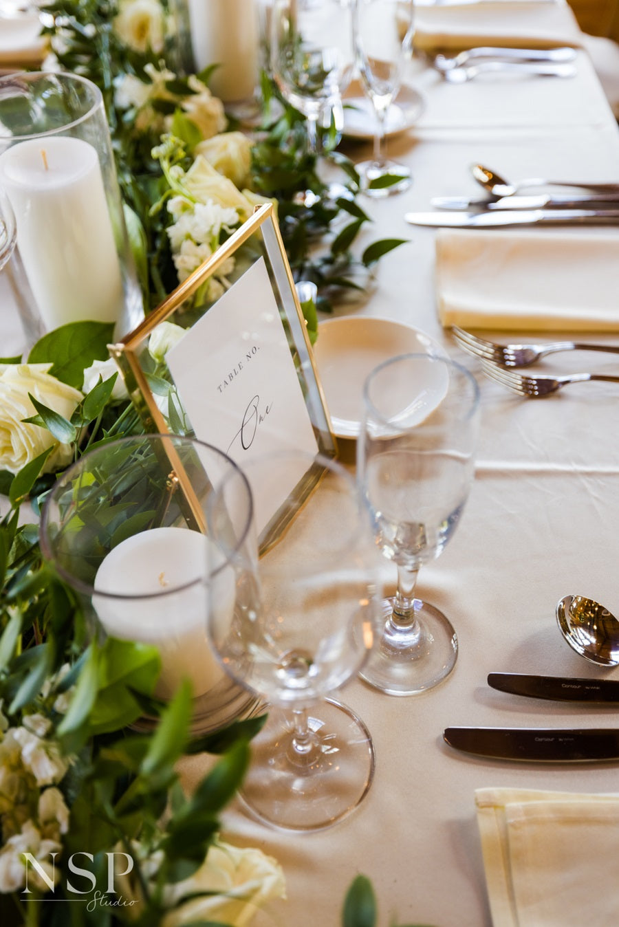Close up on an individual table setting. Loose greenery/florals with a framed table number, pillar candles, and tableware on a white table cloth.