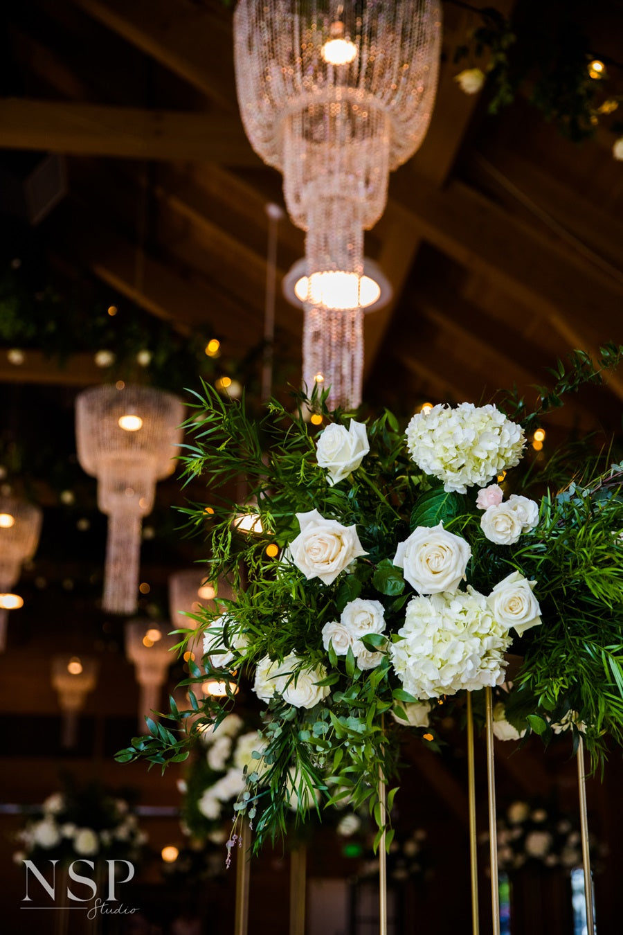 Elevated centerpieces full of greens and white florals. Background shows hanging chandeliers.