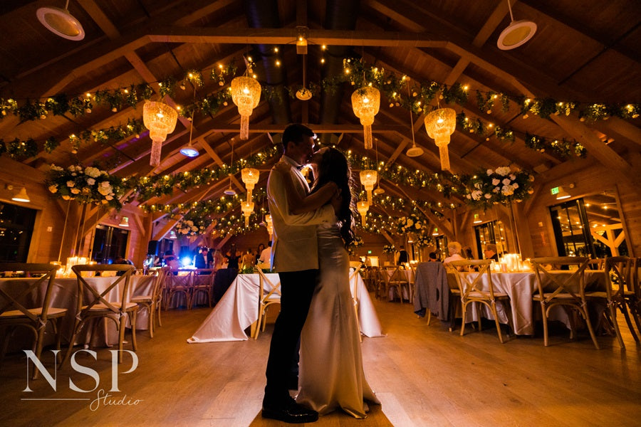 Bride and groom embrace on the dance floor, reception tables in the background. Lighting from the chandeliers and string lights glows in the background, creating a stunning atmosphere.